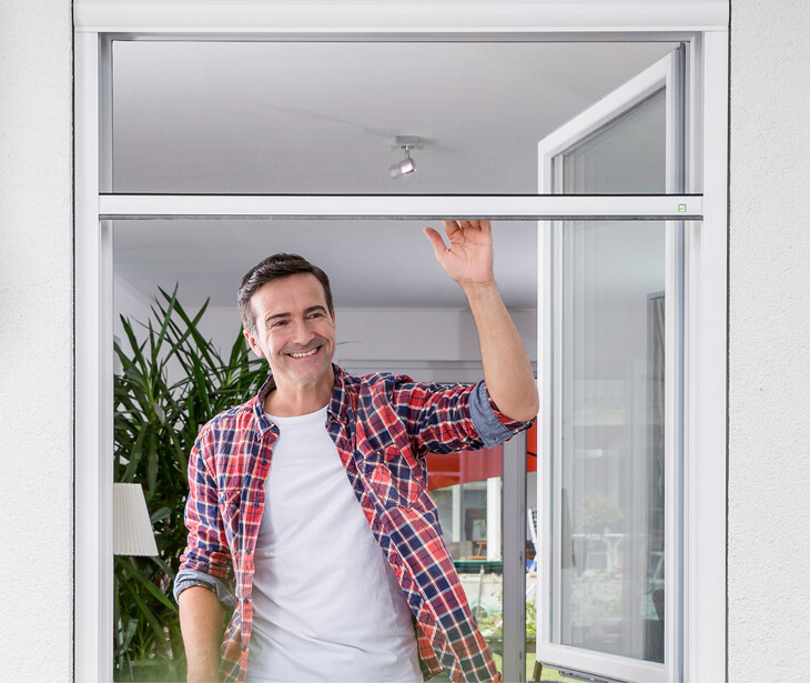 Mann in kariertem Hemd und weißem T-Shirt öffnet ein Fenster mit Insektenschutzgitter in einem hellen Raum mit Pflanzen und Möbeln.