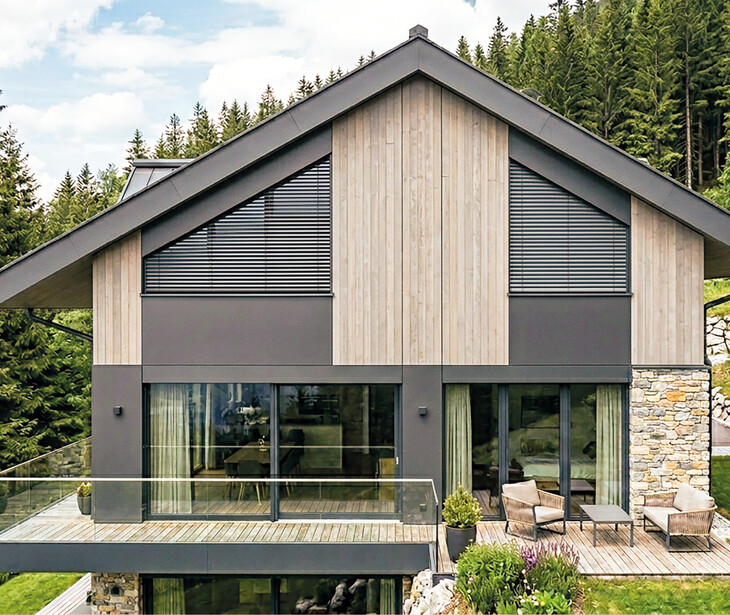 Modernes Haus mit Holz- und Steinelementen, Fenster mit Schrägraffstores, großem Balkon und Glasgeländer vor Nadelwald.