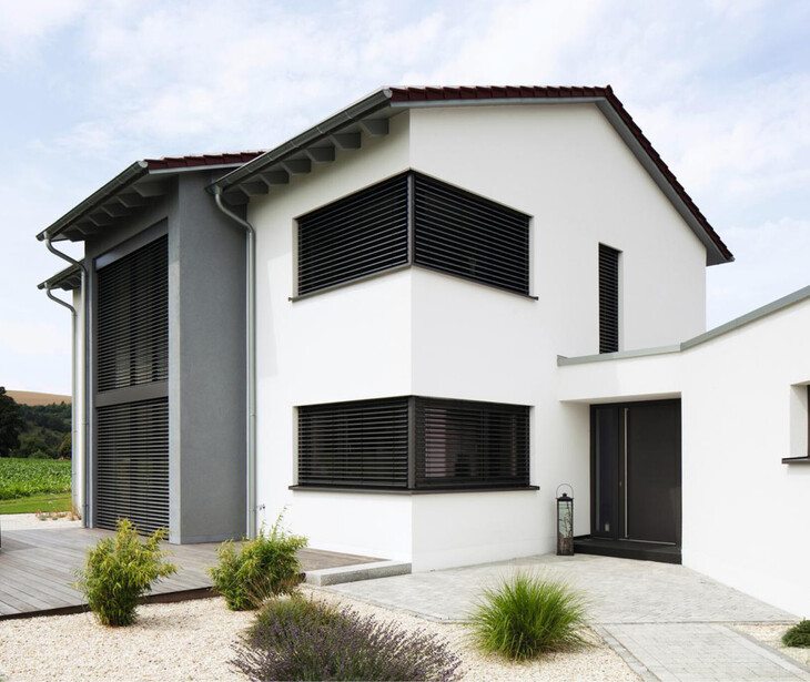Modernes, weißes Wohnhaus mit dunklen Fensterläden und Holzterrasse in ländlicher Umgebung.