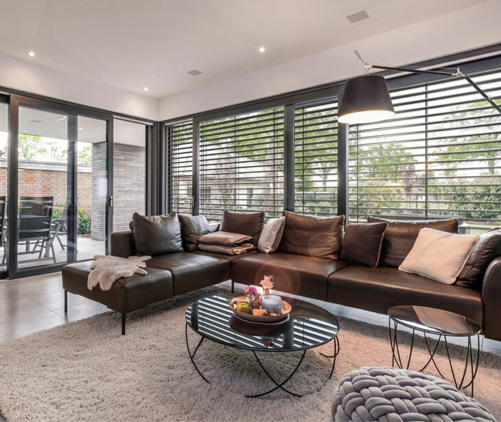 Wohnzimmer mit braunem Ledersofa, rundem Glastisch, großem Fenster mit horizontalen Jalousien und Zugang zu Terrasse.