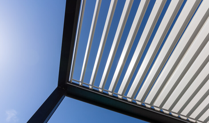 Blick nach oben auf eine moderne Pergola mit waagerechten Lamellen vor blauem Himmel.