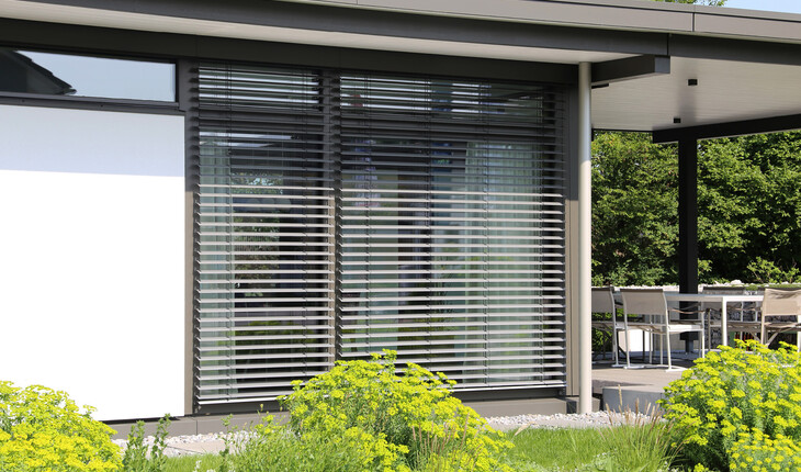 Moderner Gebäudeflügel mit großen Fenstern und Raffstores und Außenbereich mit Garten und Sitzmöbeln.