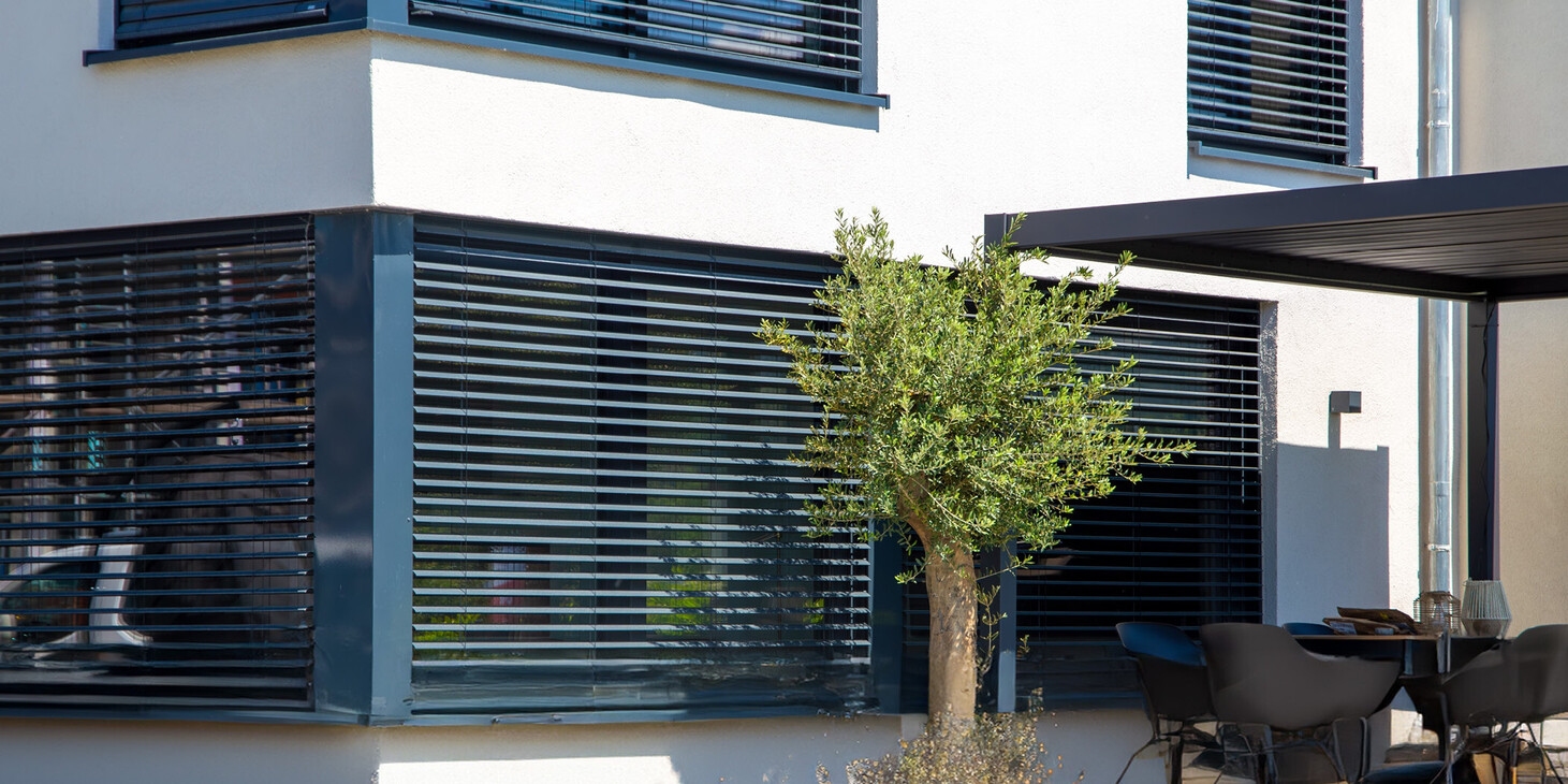 Moderner Gebäudeeck mit großen Fenstern und Jalousien, davor ein Baum und eine Terrasse mit Stühlen und Tisch unter einem Sonnenschutz.