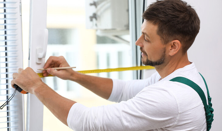 Mann mit kurz geschnittenem Haar misst mit Maßband und Bleistift an einem weißen Fensterrahmen