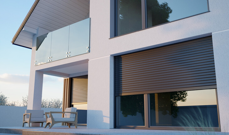 Moderner weißer Wohnhaus-Außenbereich mit Glasbalkon, zwei Stühlen und Tisch auf der Terrasse sowie geschlossenen Rollläden an großen Fenstern.