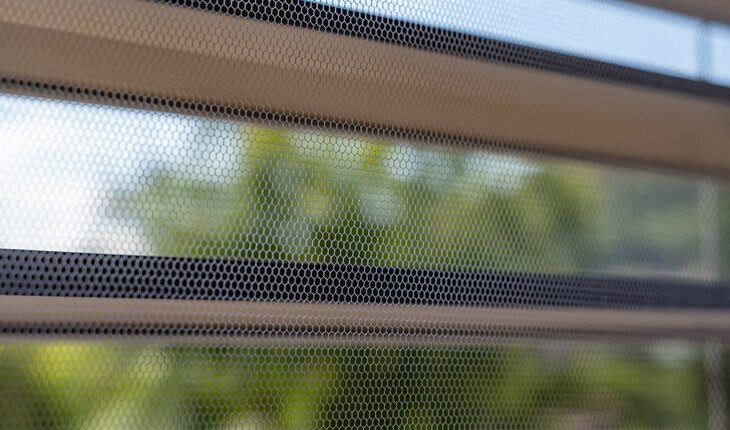 Nahaufnahme eines Fenstergitters bzw. Insektenschutz vor unscharfem Hintergrund aus Bäumen und Himmel.
