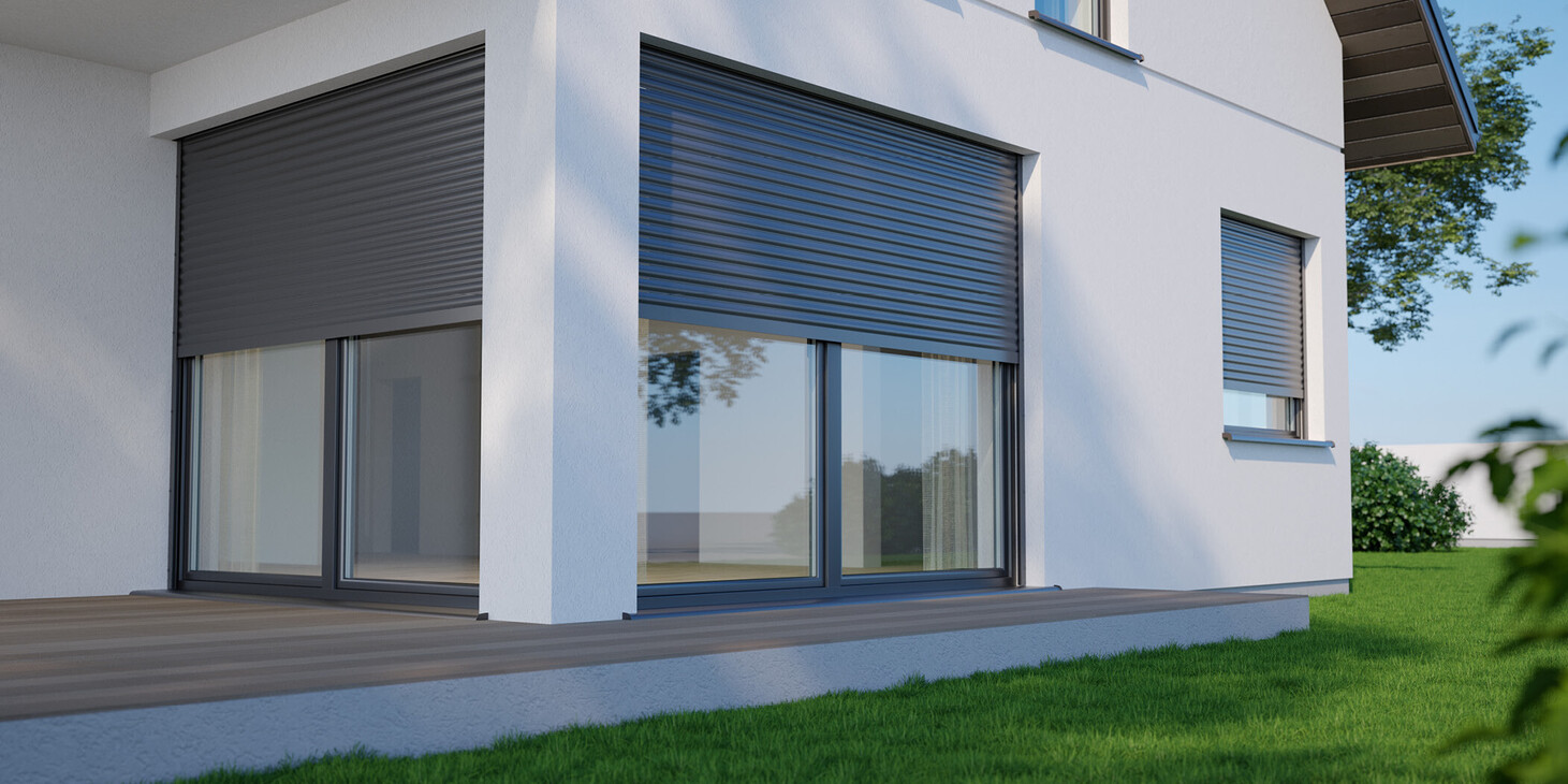 Eckansicht eines weißen Hauses mit geschlossenen grauen Rollläden an zwei Fenstern und einer Terrasse mit Holzbelag.