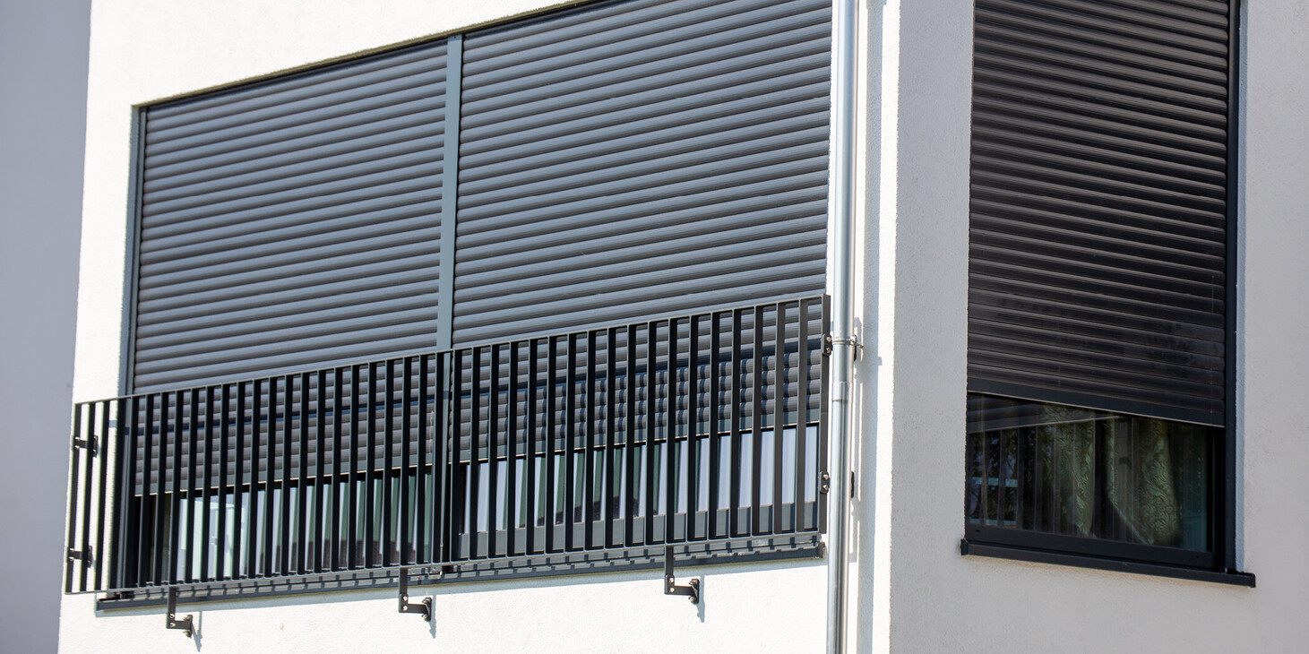Fassade eines weißen Gebäudes mit zwei großen Fenstern, die mit geschlossenen Jalousien und einem schwarzen Geländer davor versehen sind.