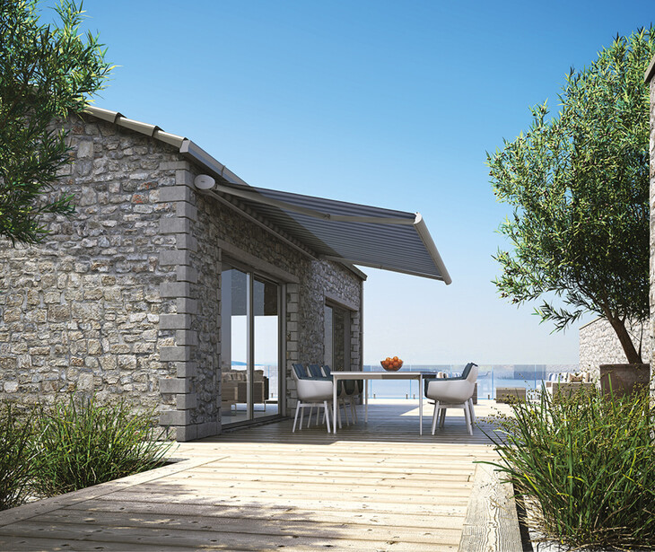 Steinernes Haus mit Sonnenschutzmarkiese, Terrasse, Tisch und Stühlen, umgeben von Pflanzen und mit Blick auf das Meer.