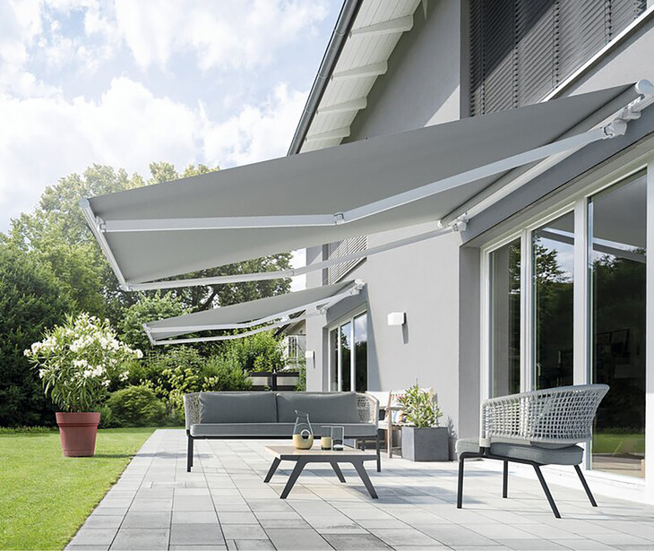Terrasse mit grauer Markise, grauem Sofa, Stuhl und Tisch vor einem modernen Haus mit großen Fenstern.