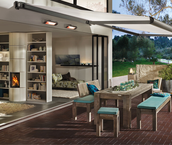 Terrasse mit großem Holztisch, gepolsterten Sitzbänken und Stühlen überdacht mit einer Sonnenschutzmarkiese vor einem modernen Wohnzimmer mit Kamin und Bücherregal.