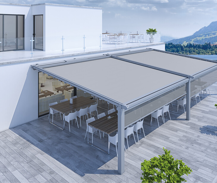 Terrasse mit zwei großen, ausfahrbaren Markisen über Tischen und Stühlen vor einem modernen Gebäude mit Glasgeländer und Bergblick.