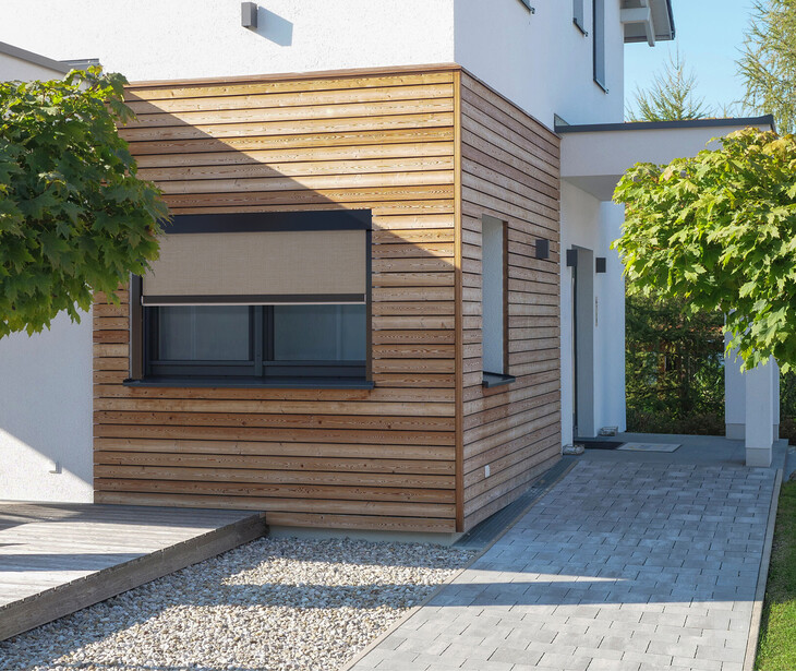Eingangsbereich eines modernen Hauses mit Holzverkleidung, Fenster mit Schonneschutzmarkiese, zwei kleinen Bäumen und gepflastertem Weg.