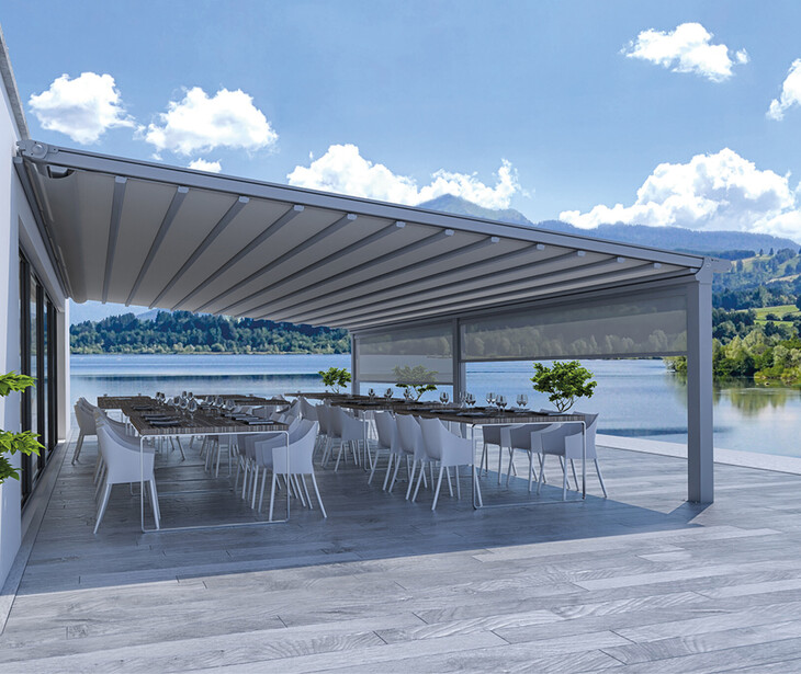 Terrasse mit moderner Pergola, mehreren Tischen und Stühlen vor einem See und bewaldeten Hügeln.