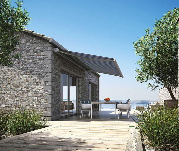 Casa in pietra con tenda parasole, terrazza, tavolo e sedie, circondata da piante e affacciata sul mare.
