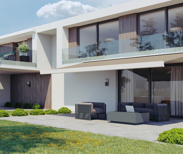 Moderner zweigeschossiger Wohnbau mit großer Glasfront, Terrasse mit Sitzmöbeln und Markise, umgeben von Rasen und kleinen Sträuchern.