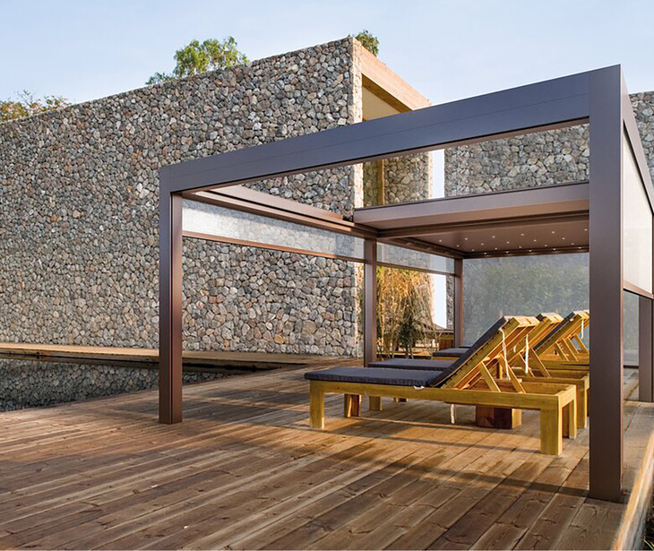 Moderner Holzsteg mit Sonnenschutzrahmen und Liegestühlen vor einer Natursteinmauer.