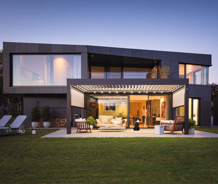 Modernes zweigeschossiges Haus mit großer Glasfront und überdachter Terrasse auf gepflegtem Rasen.