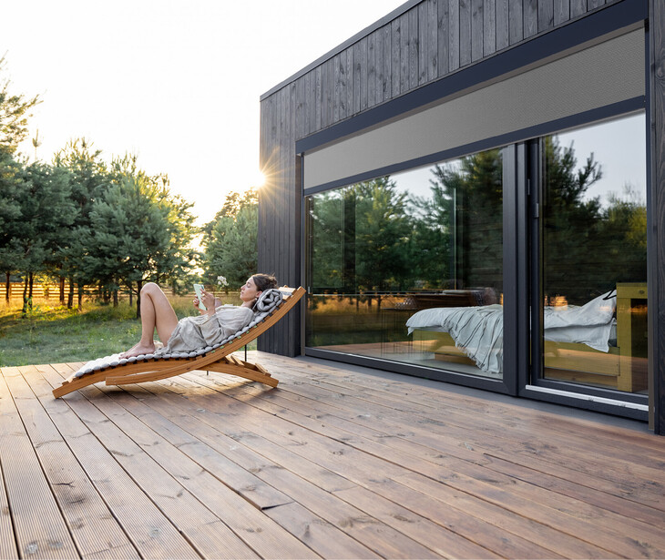 Person liegt auf einer Holzliege auf einer großen Holzterrasse neben einem modernen Haus mit großen Fenstern und Sonnenschutzmarkiesen.