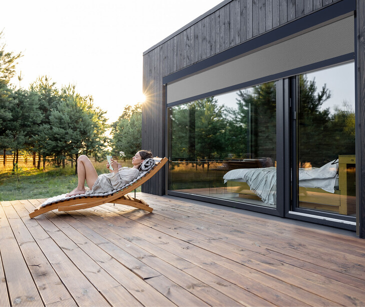 Una persona è sdraiata su una sdraio di legno su un'ampia terrazza in legno accanto a una casa moderna con grandi finestre e tende da sole.