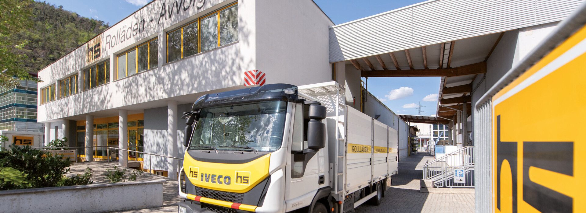 Lkw mit Aufschrift hs iveco hs vor einem weißen Gebäude mit gelben Fensterrahmen und der Aufschrift Rolladen Avvolgibili.