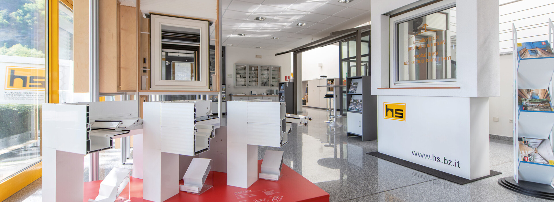 Innenraum eines modernen Ausstellungsraums mit weißen Modellen auf rotem Podest und großen Fenstern.